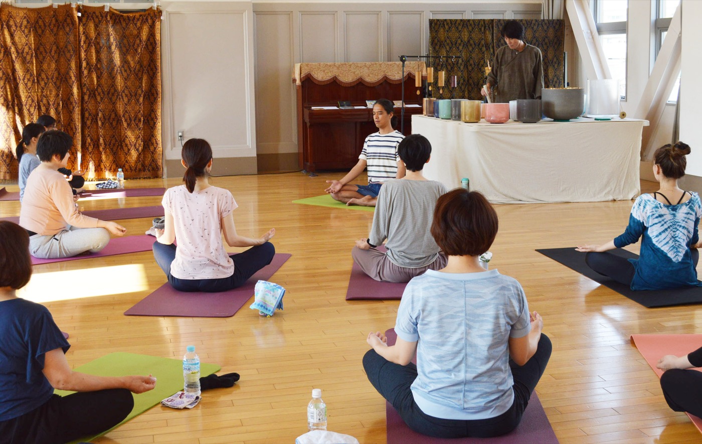 鴨江アートセンターの会場で輪になって座り、クリスタルボウルの音に耳を傾ける参加者たち、奥には色とりどりのボウルが並ぶ演奏台、やわらかな室内の光